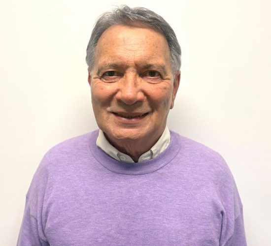 Portrait of an older man smiling, wearing a lavender sweater over a light collared shirt against a plain white background.