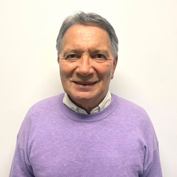 Portrait of an older man smiling, wearing a lavender sweater over a light collared shirt against a plain white background.
