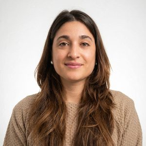 Portrait of a woman with long brown hair, wearing a beige knit sweater, looking at the camera with a subtle smile.