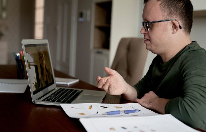 Man wearing glasses in a green shirt talks on a video call, gesturing at a laptop on a desk with papers nearby.