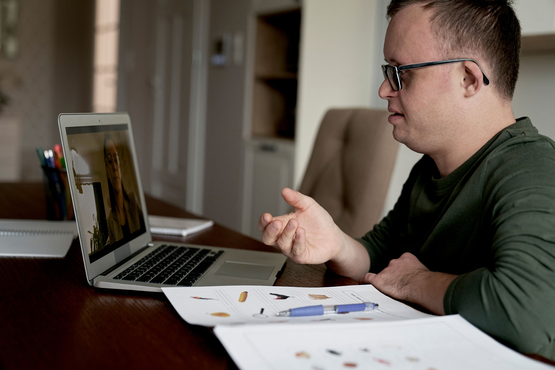 Man wearing glasses in a green shirt talks on a video call, gesturing at a laptop on a desk with papers nearby.