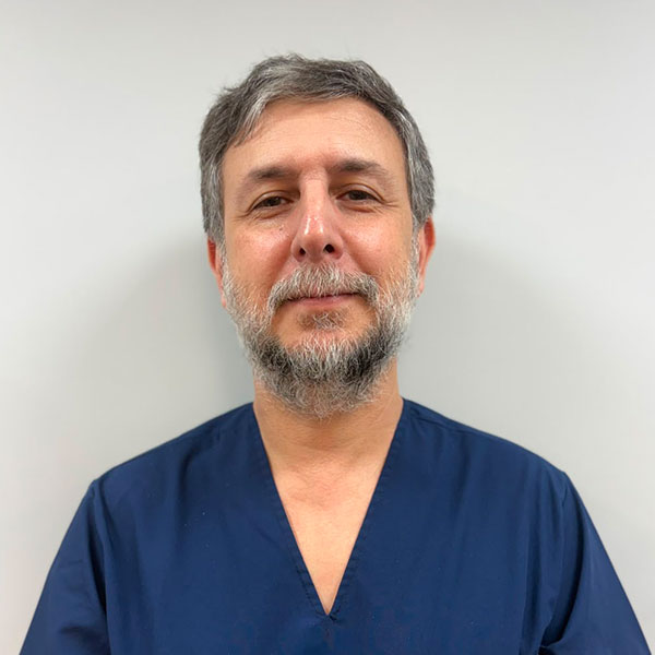 Portrait of a middle-aged man with gray hair and a gray beard, wearing navy scrubs against a light gray background, looking at the camera with a calm expression, suggesting a healthcare professional.