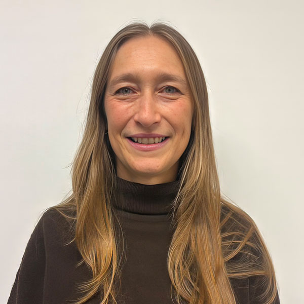 Smiling woman with long blond hair wearing a dark brown turtleneck against a light background.