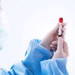 Medical professional in blue gown and gloves holding a small vial with a red cap