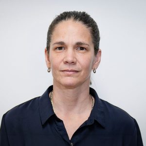 Portrait of a woman with dark hair tied back, wearing a navy button-down shirt, with a neutral expression against a light background.