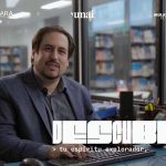 Portrait of a man in a blazer seated at a desk typing on a keyboard in an office with bookshelves in the background and logos on the glass behind him AZARA umai Mulata Films