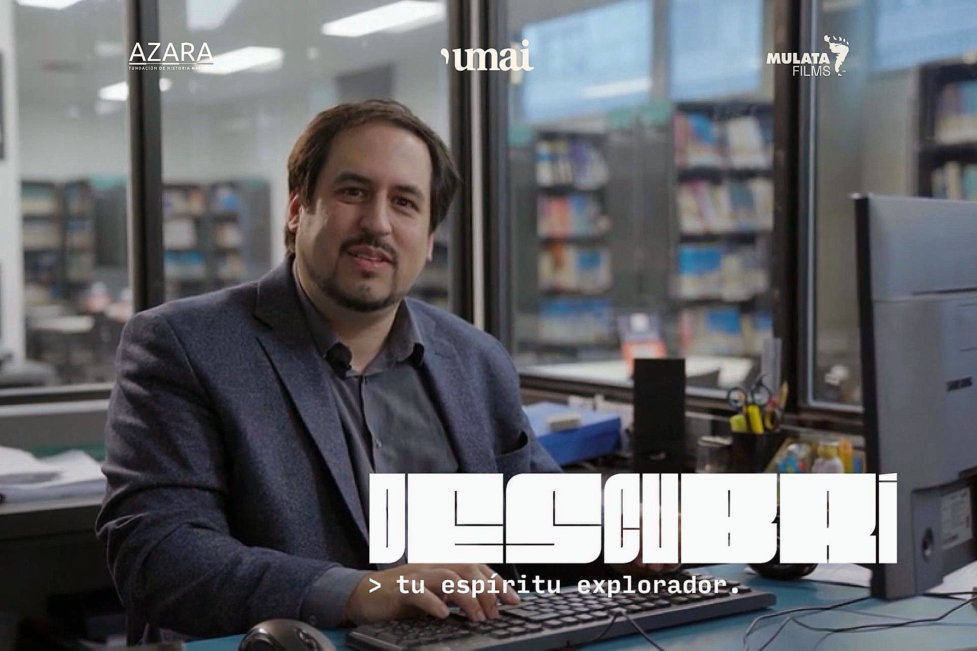 Portrait of a man in a blazer seated at a desk, typing on a keyboard in an office with bookshelves in the background and logos on the glass behind him (AZARA, ’umai’, Mulata Films).
