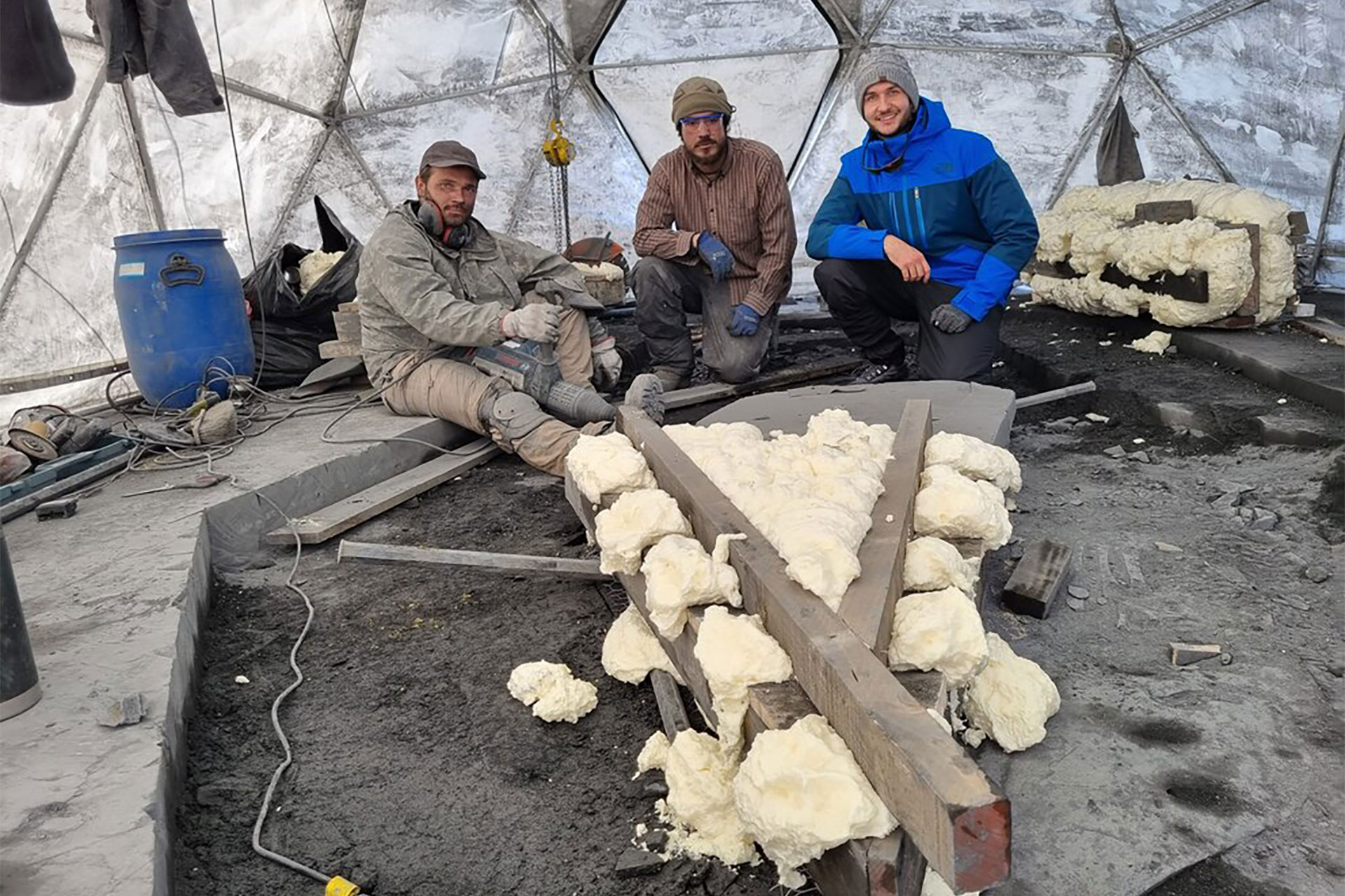 Three construction workers inside a geodesic dome, sitting among foam insulation and scattered tools.