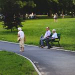 An elderly man and woman sit on a green bench by a paved path in a grassy park while a woman in a floral skirt walks past and other people relax in the background