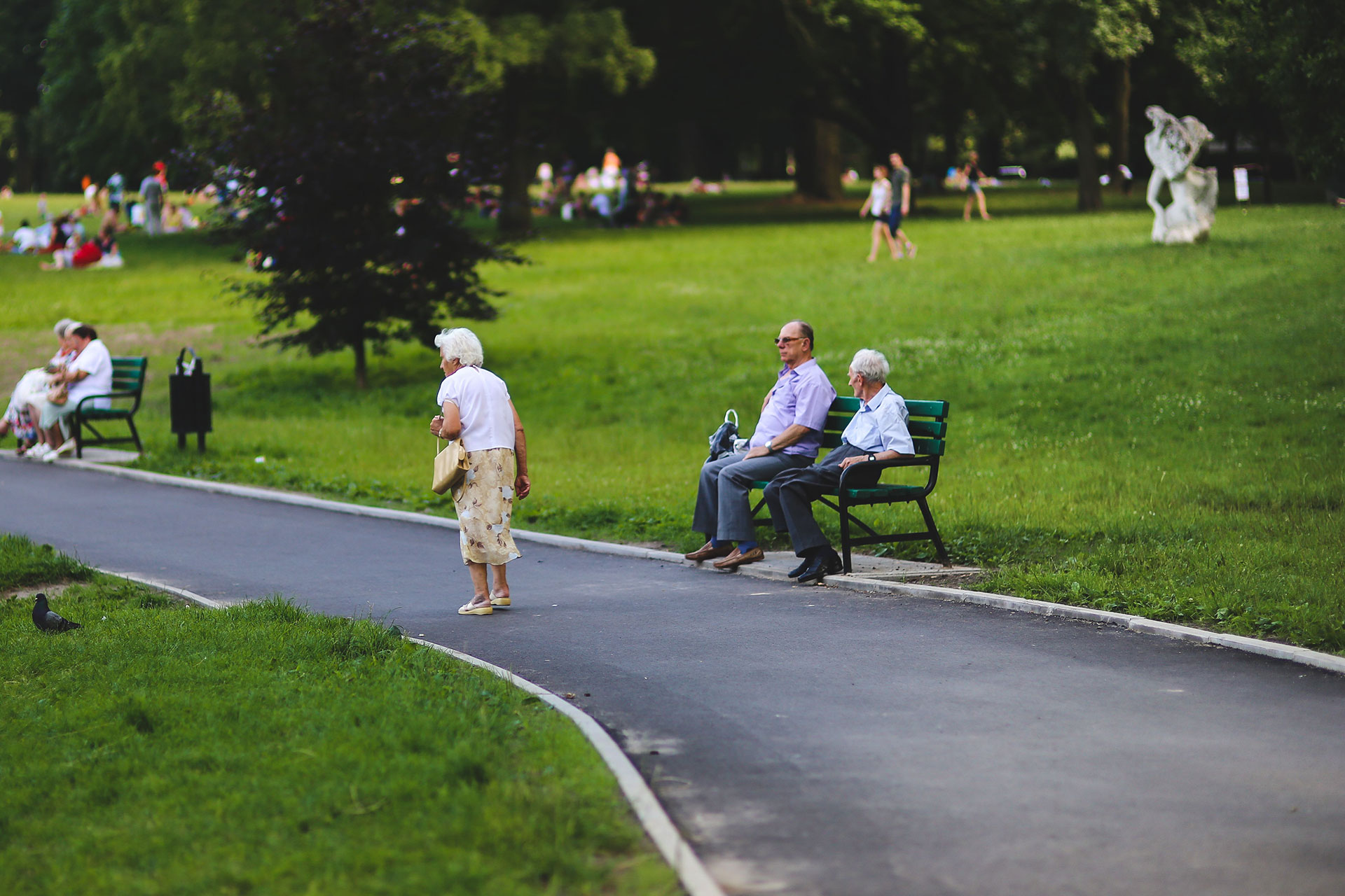 An elderly man and woman sit on a green bench by a paved path in a grassy park while a woman in a floral skirt walks past and other people relax in the background.