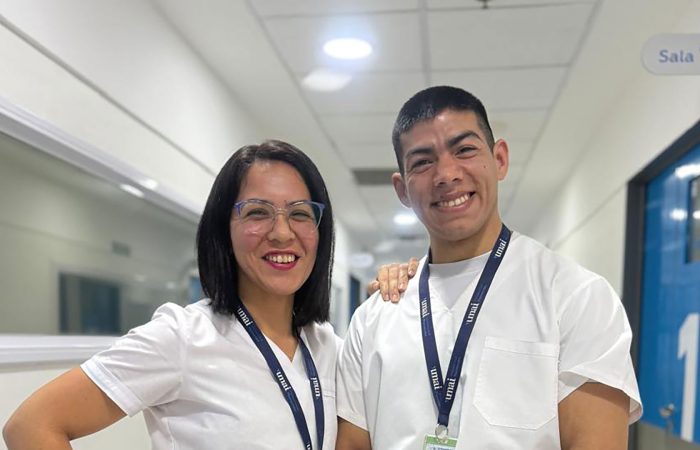 Two healthcare workers in white scrubs smiling in a hospital hallway with their arms around each other.