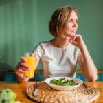 Woman at a table holding a glass of orange juice with a salad bowl in front and a green wall behind her