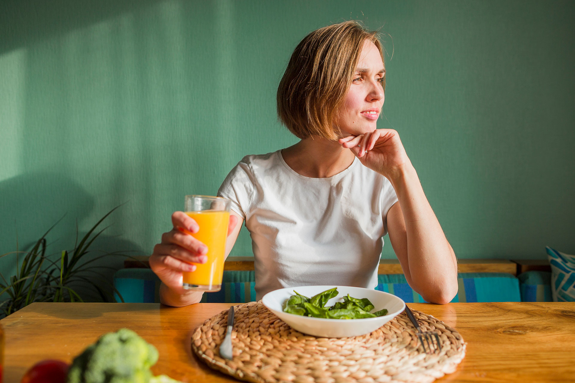 Woman at a table holding a glass of orange juice, with a salad bowl in front and a green wall behind her.