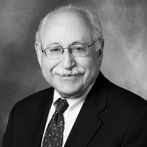 Black-and-white portrait of an older man with glasses, a white mustache, wearing a dark suit and patterned tie, smiling in a professional headshot.