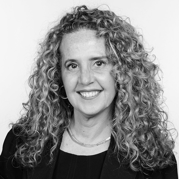 Professional headshot of a woman with long curly hair smiling at the camera, wearing a dark blazer, on a light background.