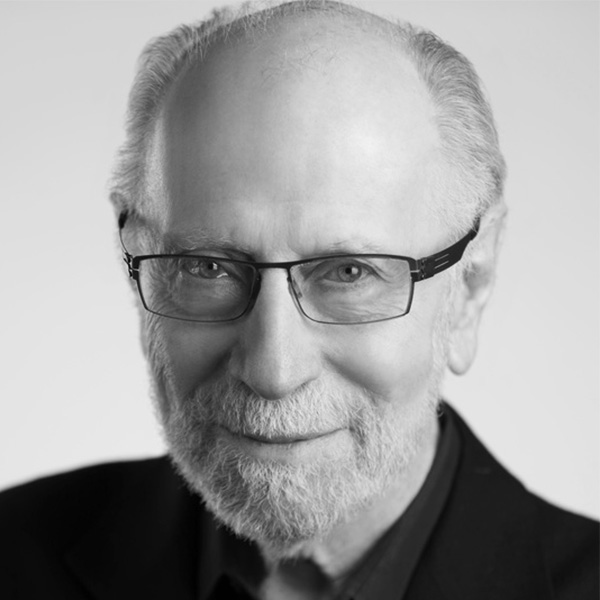 Close-up black-and-white headshot of an older man with rectangular glasses, a trimmed white beard, and a gentle smile, wearing a dark jacket.