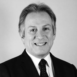 Professional headshot of a smiling man in a dark suit and polka-dot tie against a light background, portrait for corporate use.