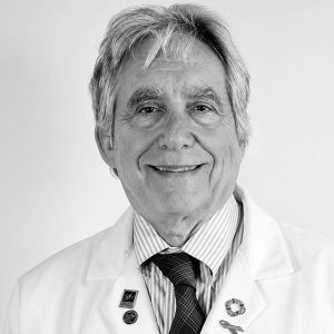 Smiling senior male doctor in a white coat with striped shirt and tie, wearing medical pins, against a neutral background