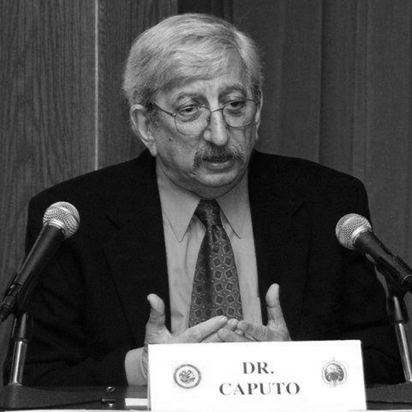 Older man in a suit speaking at a panel with two microphones; nameplate reads Dr. Caputo.