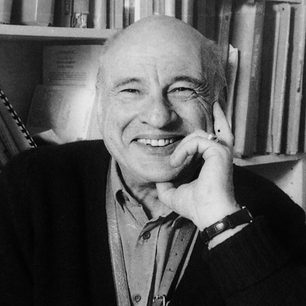 Smiling elderly man talking on a landline phone, seated beside a bookshelf.