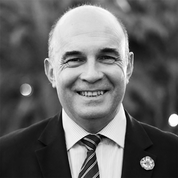 Smiling man in a dark suit and striped tie, outdoors, wearing a lapel pin.