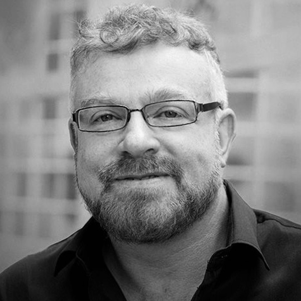 Portrait of a smiling man with short, gray hair and a beard, wearing glasses and a dark shirt; grayscale photo.
