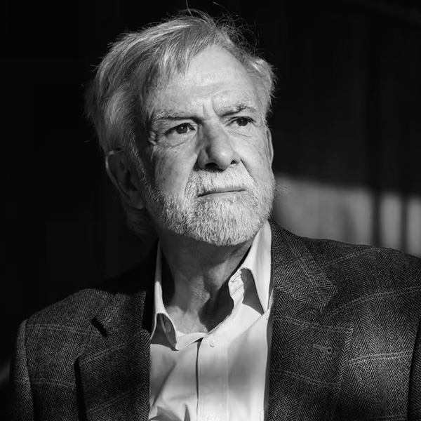 Older man with gray hair and beard wearing a blazer, looking pensively to the side.