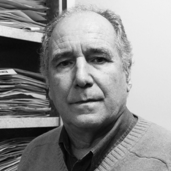 Black-and-white portrait of a middle-aged man in a sweater, standing beside a filing shelf with stacked folders.