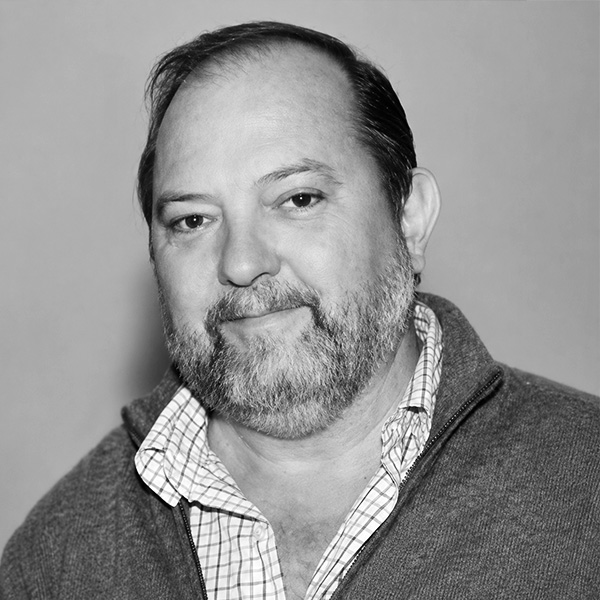 Portrait of a middle-aged man with a short beard, wearing a checkered shirt and a zip-up sweater, looking at the camera with a gentle smile.