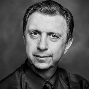 Black-and-white head-and-shoulders portrait of a middle-aged man in a dark shirt and tie, looking at the camera with a subtle smile.