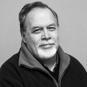Portrait of a smiling middle-aged man with gray beard and short hair, wearing a dark fleece jacket against a plain background.