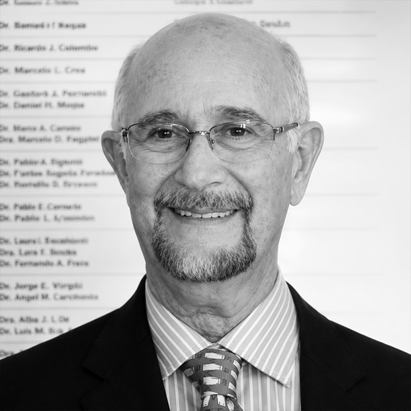 Professional head-and-shoulders portrait of an older man with glasses, a goatee, and a suit, standing in front of a wall of names.