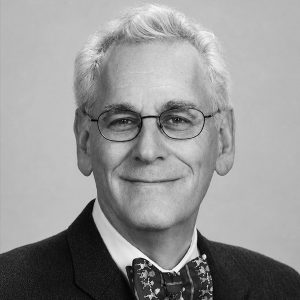 Smiling older man with gray hair and round glasses, wearing a dark suit and a patterned bow tie.