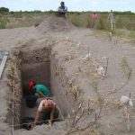People dig a deep trench in sandy dune terrain using buckets and tools with a few workers and surveying gear nearby
