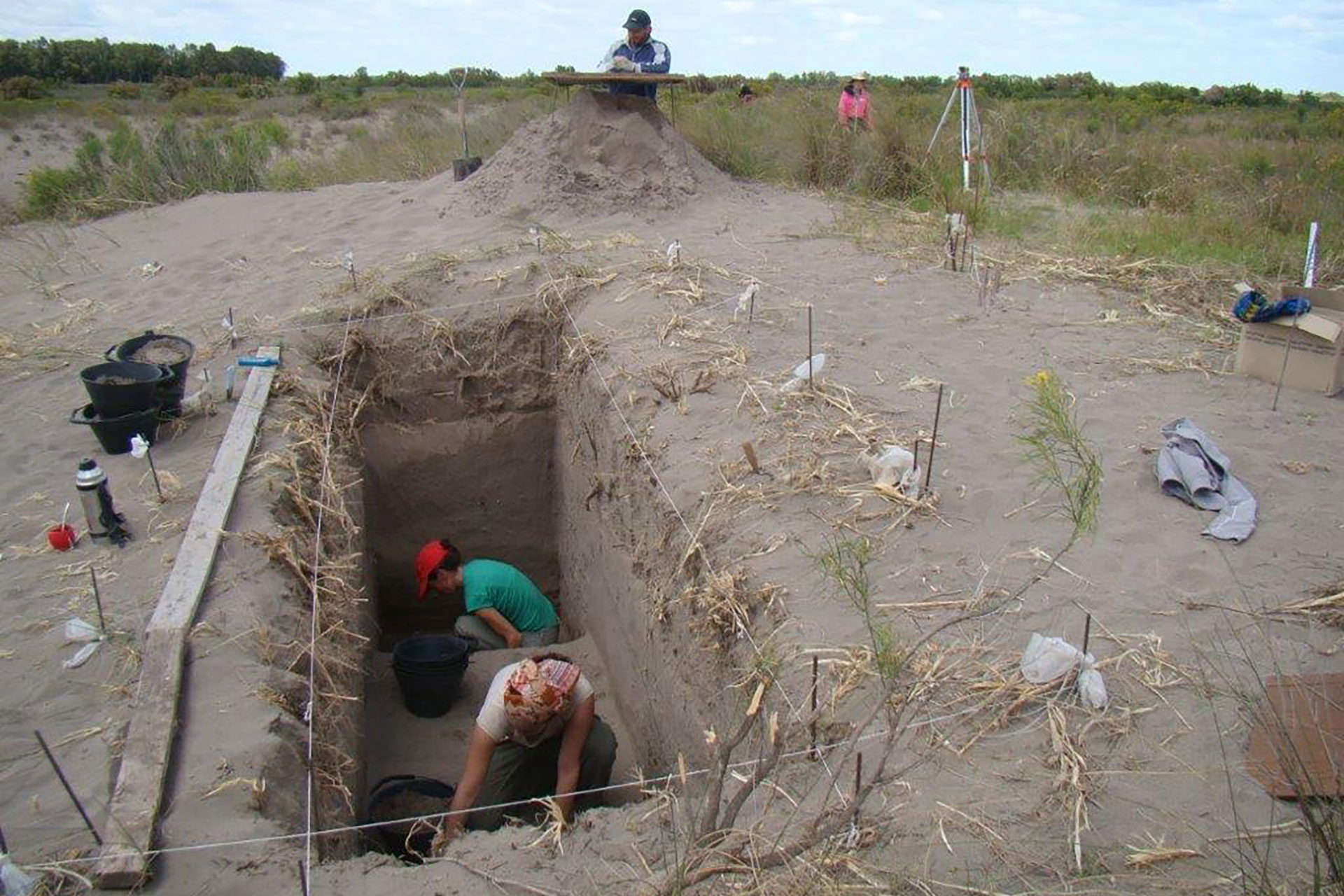 People dig a deep trench in sandy dune terrain using buckets and tools with a few workers and surveying gear nearby.