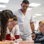 Nurse in white scrubs measures an elderly womans blood pressure at a clinic desk while others look on