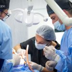 Team of surgeons in blue gowns performing an operation wearing masks and caps under bright overhead lights