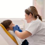 Dental assistant in a white coat with blue gloves attends to a smiling child patient seated in a dental chair with yellow headrest