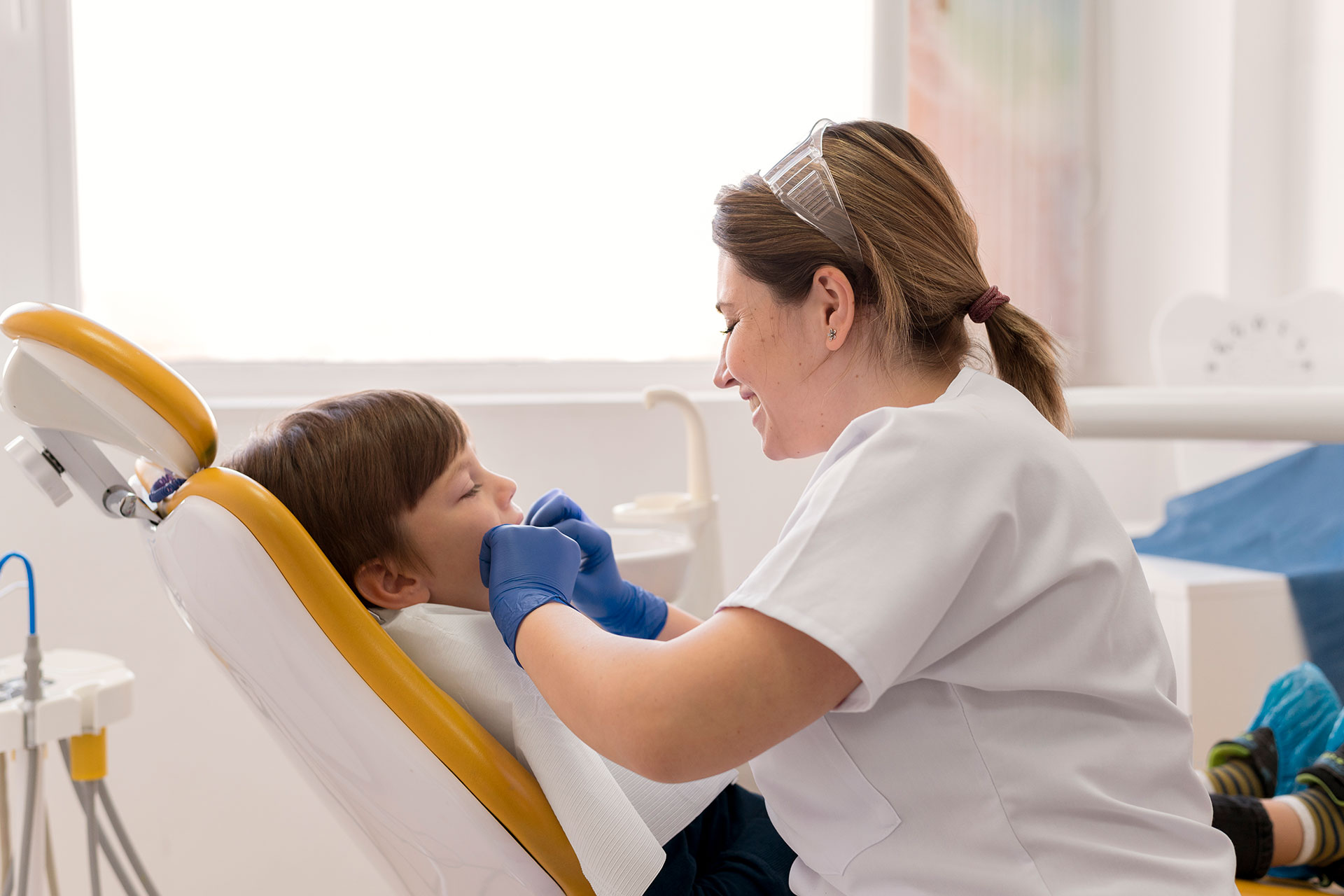 Dental assistant in a white coat with blue gloves attends to a smiling child patient seated in a dental chair with yellow headrest.