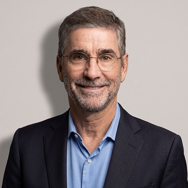 Smiling middle-aged man with gray hair and glasses wearing a navy blazer and light blue shirt in a neutral background.