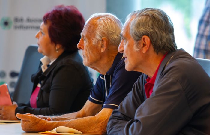 Three adults seated at a conference table in a discussion, with a woman holding an orange phone visible at left.