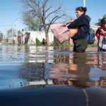 People wading through waist deep floodwater on a street carrying belongings and a child with a pole nearby