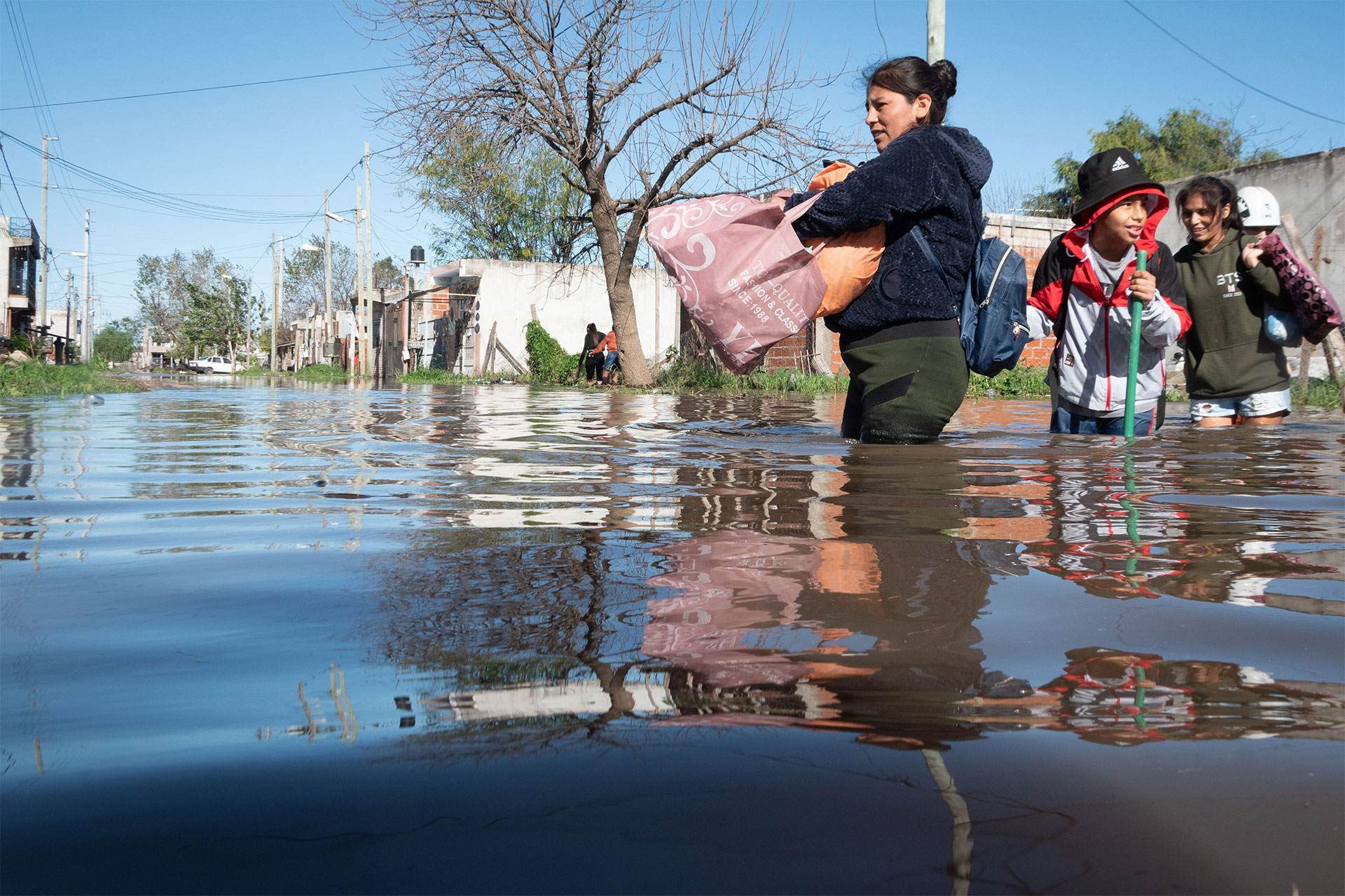 People wading through waist-deep floodwater on a street, carrying belongings and a child with a pole nearby.