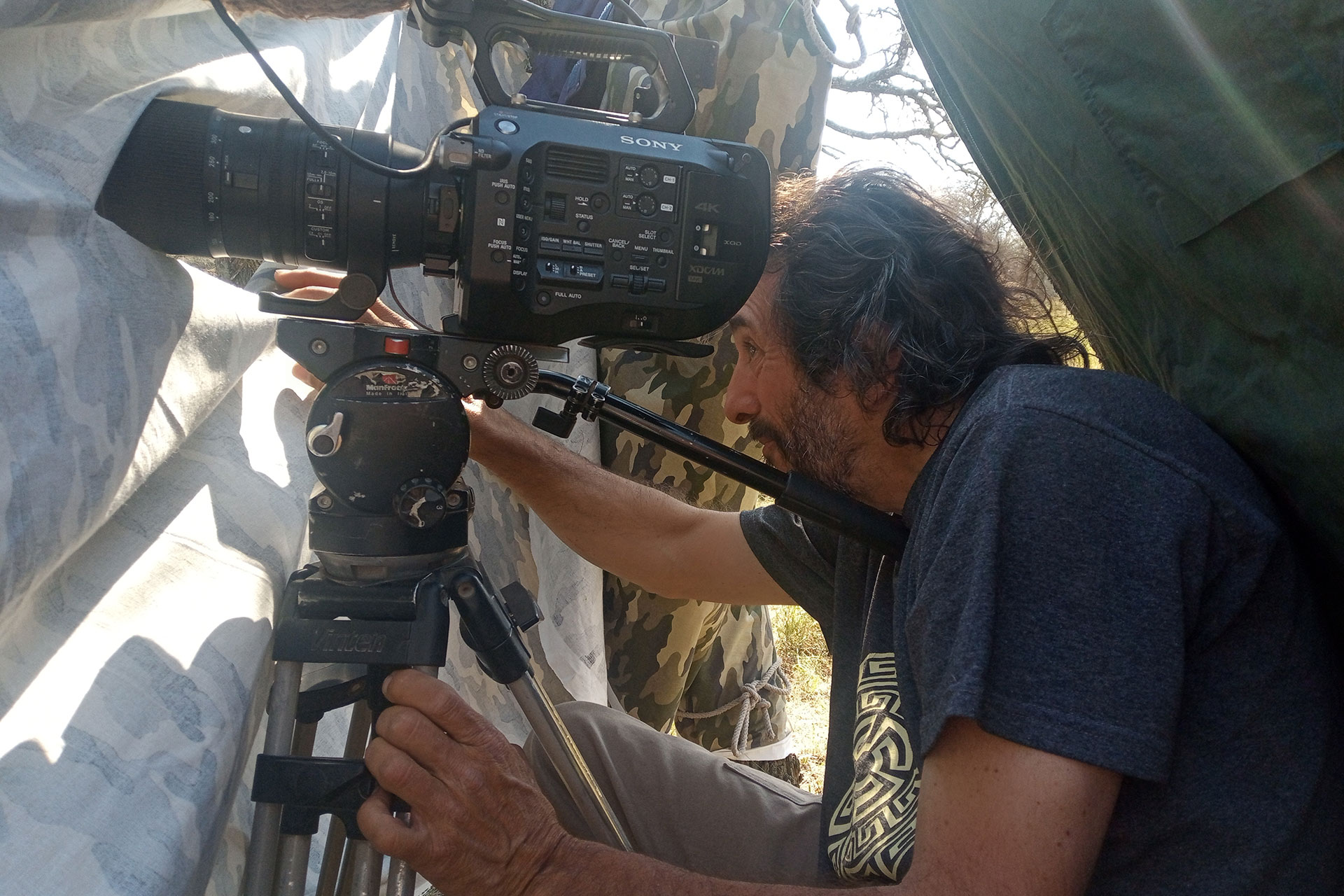 Man operating a Sony video camera mounted on a tripod inside a camouflage shelter outdoors