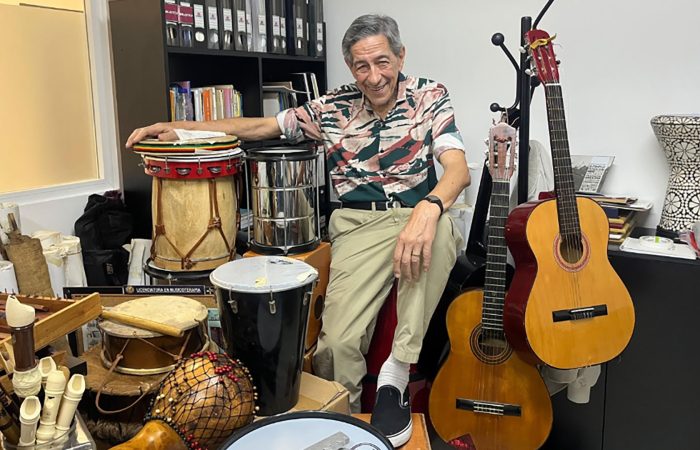 Smiling older man sits among percussion instruments (congas, bongos) and two guitars in a cluttered music room/studio.
