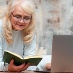 Older woman with glasses reading a green hardcover book at a desk laptop nearby