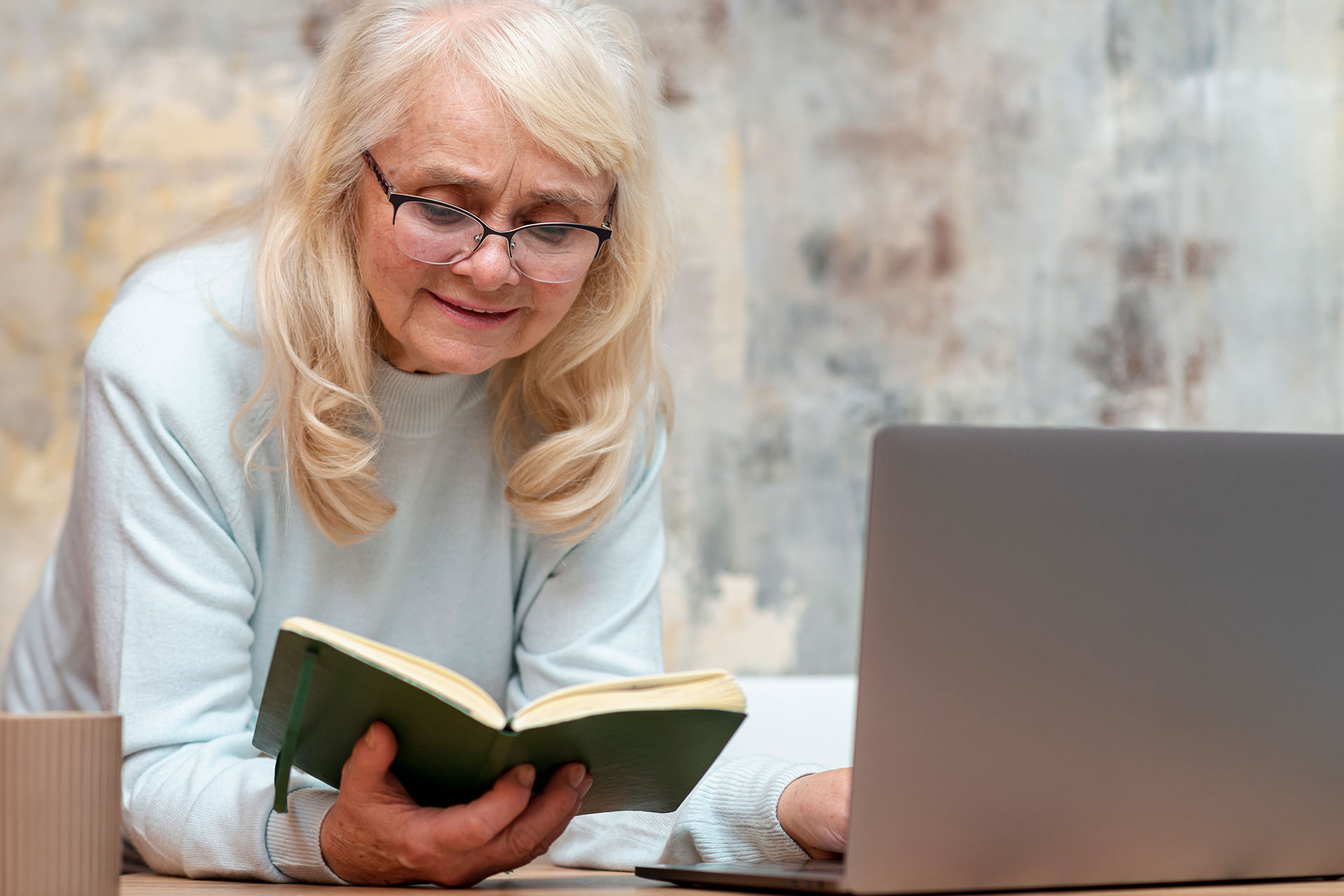 Older woman with glasses reading a green hardcover book at a desk, laptop nearby.